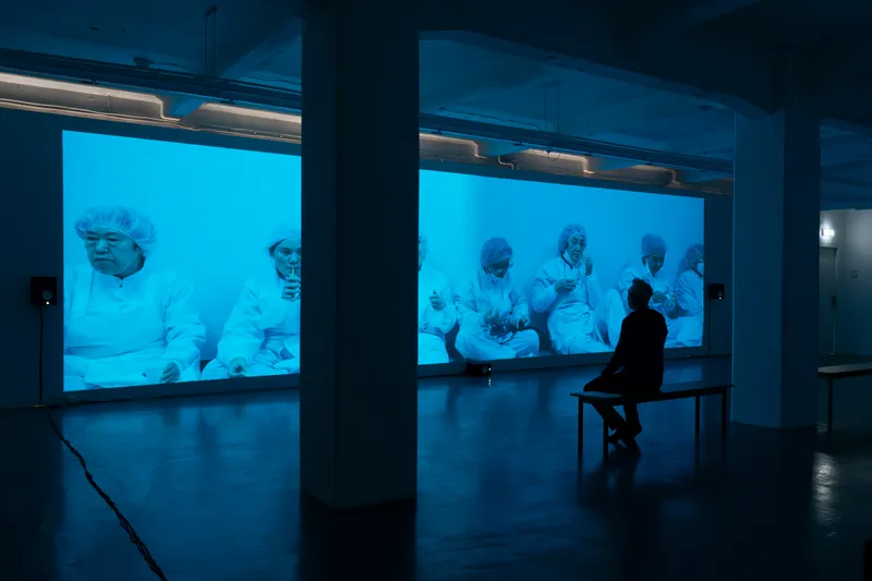 A visitor on a bench in front of an ultra-wide blue projection of factory workers in white