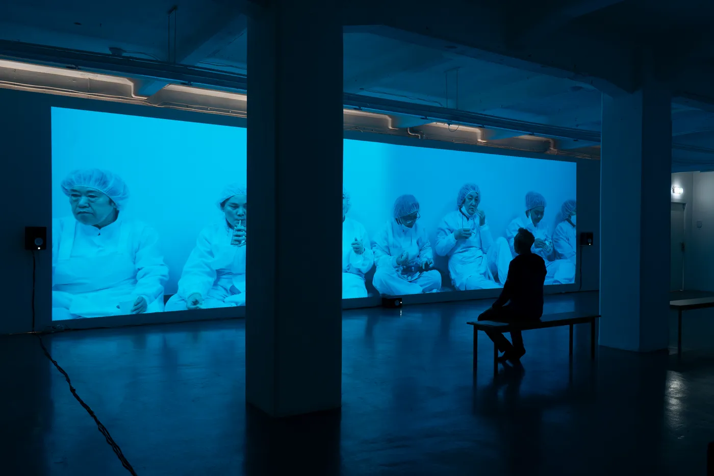 A visitor on a bench in front of an ultra-wide blue projection of factory workers in white