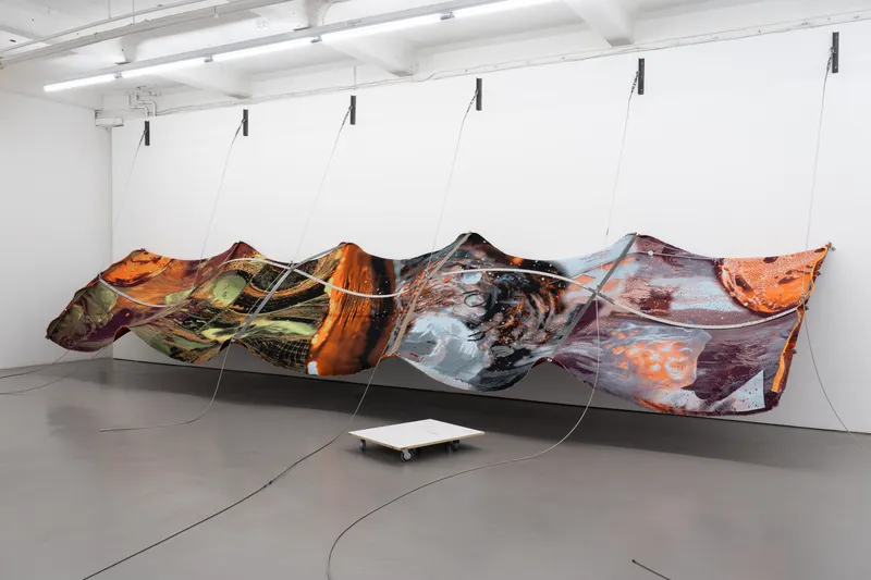 A long printed textile in orange, grey and blue hung on wires along a white wall