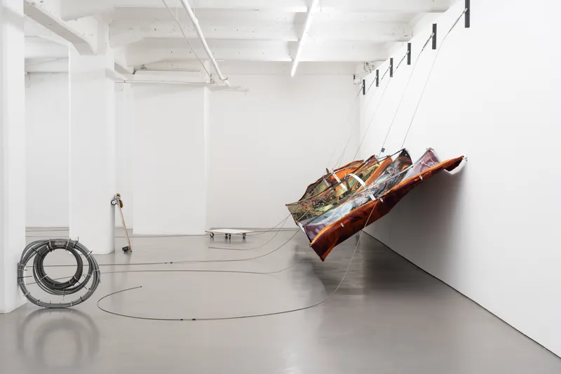 A long white gallery with a printed textile hanging diagonally on cables, coiled cables and an overturned table on the floor