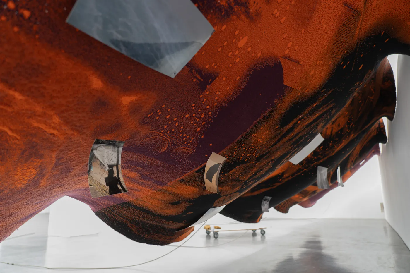 A printed textile in orange and deep red seen from below, hanging in a wave with small photographs fixed to it, a wheeled board on the floor beyond