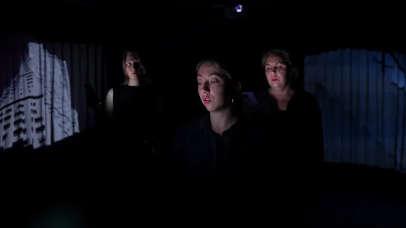 Film still: three performers in a dark room, one reading aloud, a projection of a tower block on the curtain behind