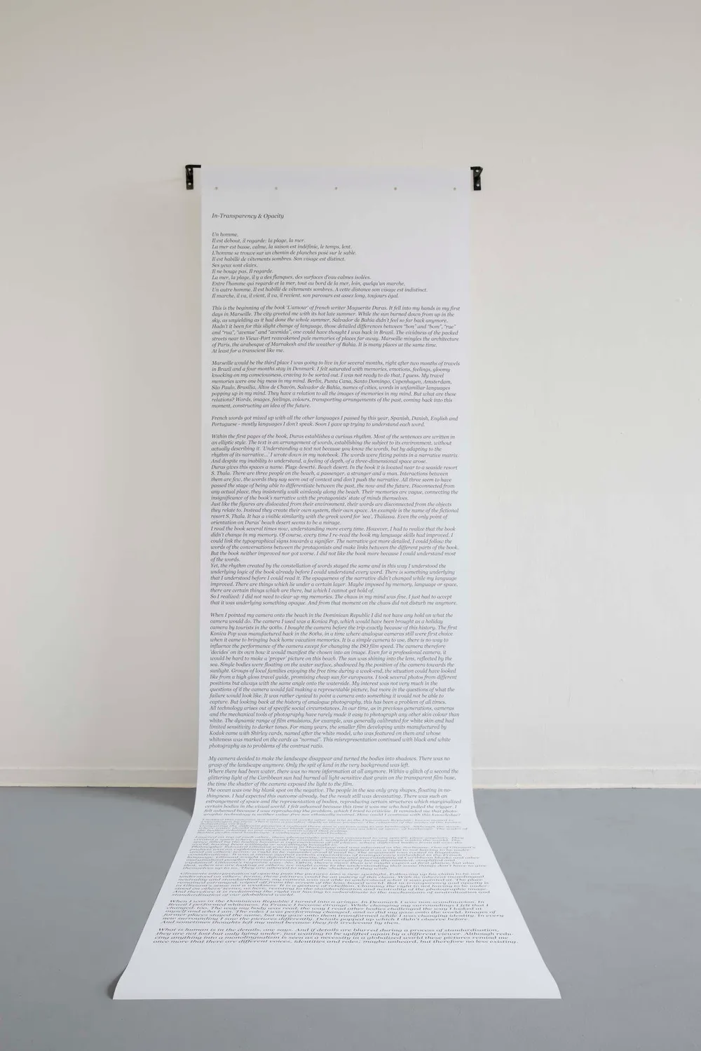 A long sheet of printed text hanging from a rod on a white wall and running onto the floor