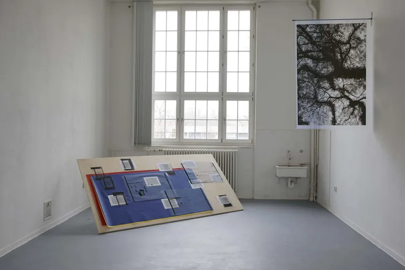 A white room with windows: a long printed text hangs from the wall and runs across the floor, blue boards lean against the far wall