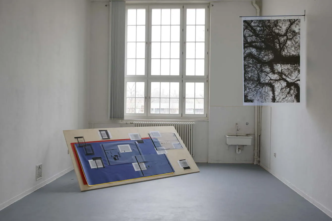 A white room with windows: a long printed text hangs from the wall and runs across the floor, blue boards lean against the far wall