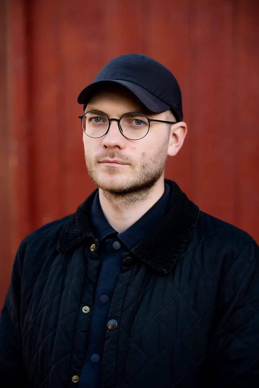 Lou Mouw in glasses and a dark cap in front of a rust-red wall