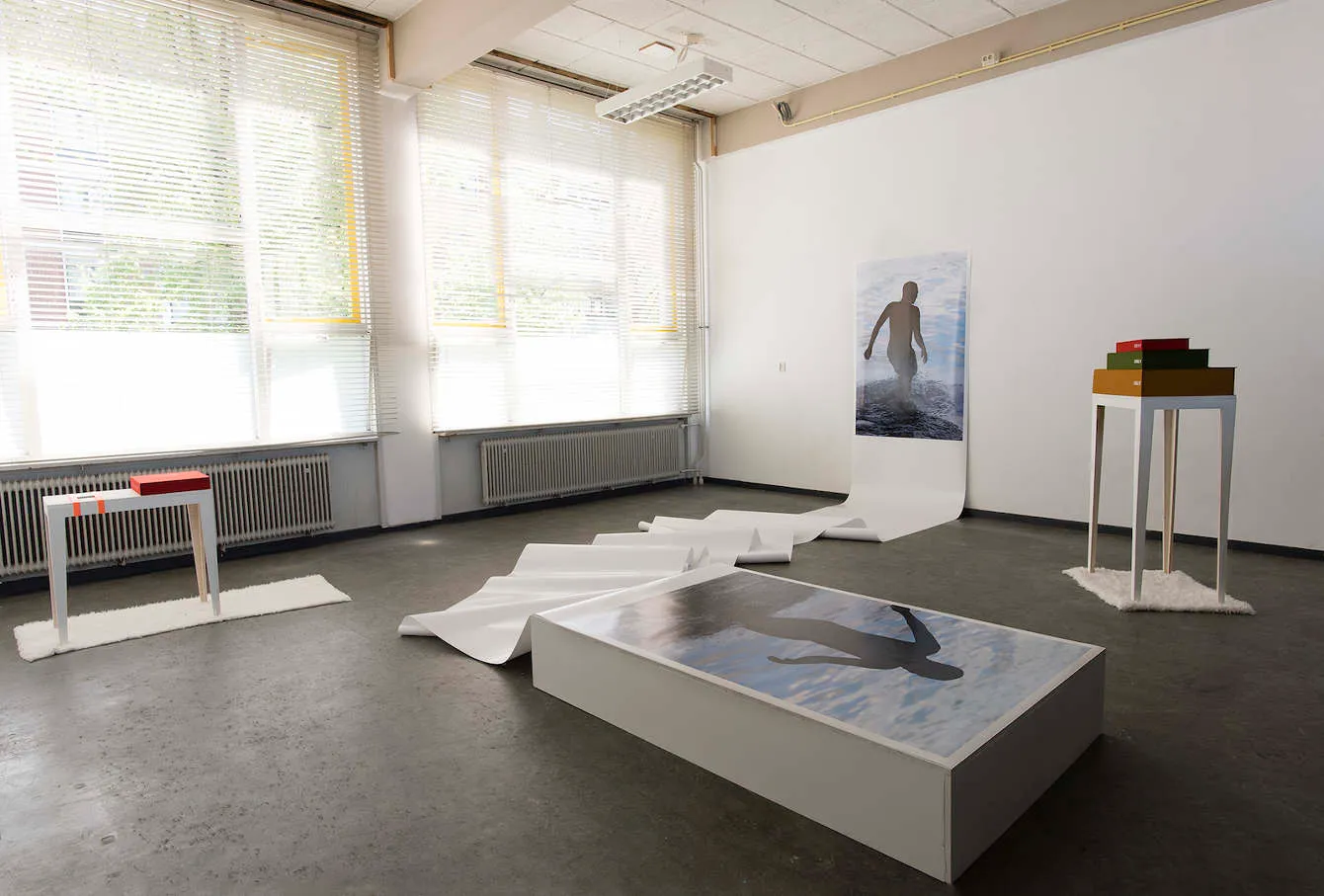 A gallery with a long paper scroll on the floor, a light box, a large photo of a swimmer and a plinth with stacked boxes