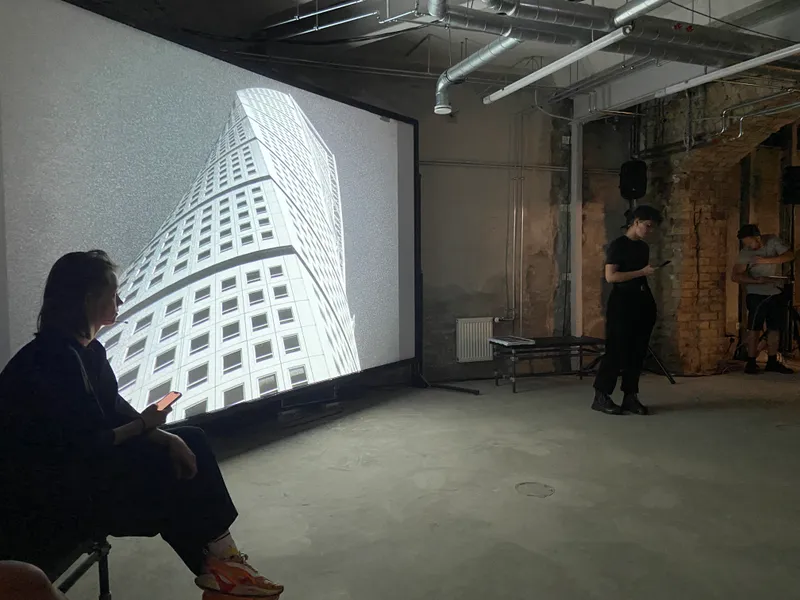 A performer sits reading in front of a large projection of Malmö's Turning Torso tower