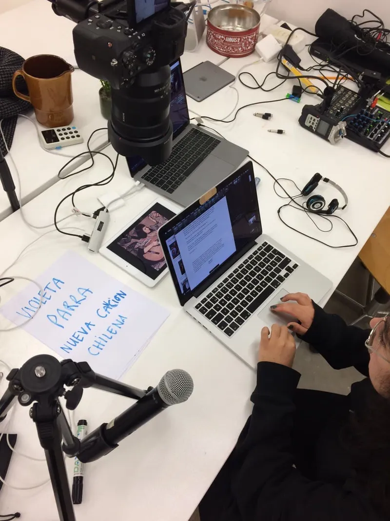 A desk with a camera, laptops, a mixer, a microphone and a sheet reading Violeta Parra, Nueva Canción Chilena