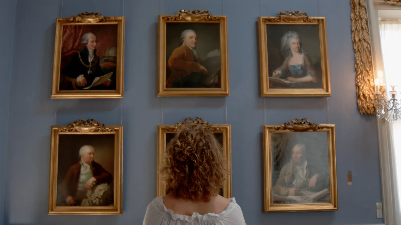 A woman with curly hair seen from behind, looking at six portraits of men and women in gilded frames on a blue wall