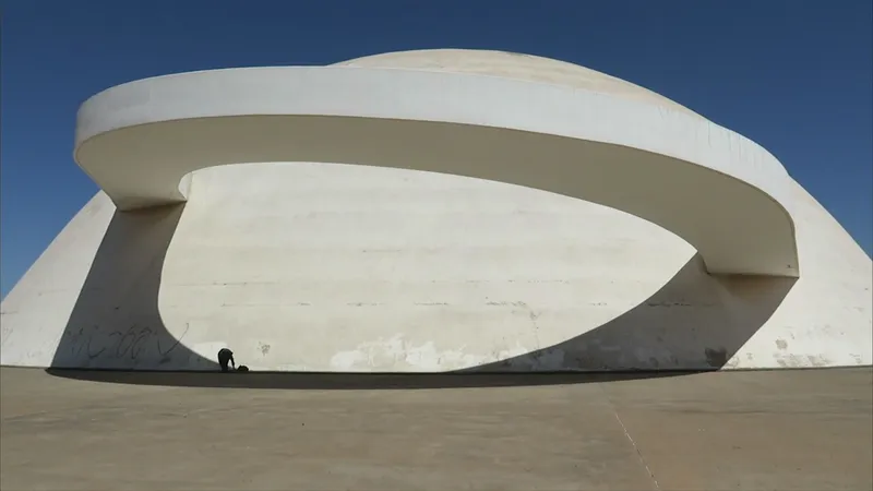 A person crouches in the shadow of the curved concrete ring of the National Museum in Brasília