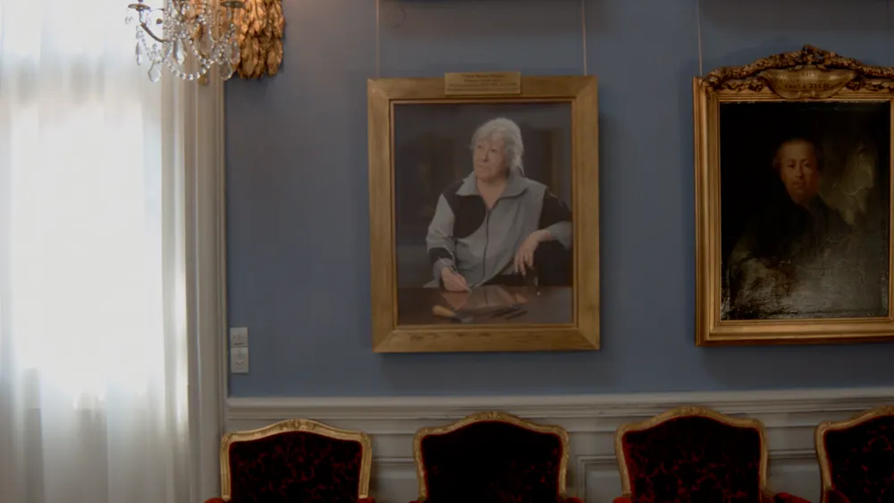A painted portrait of an older woman in a grey jacket on a blue wall, chairs below
