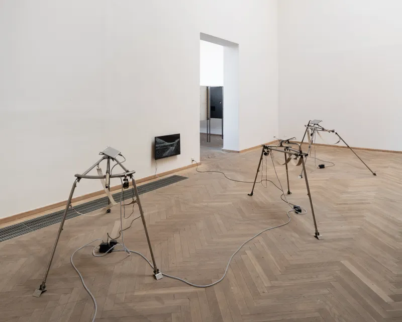Two steel tripods with small screens in a gallery with a herringbone floor, a third screen on the wall