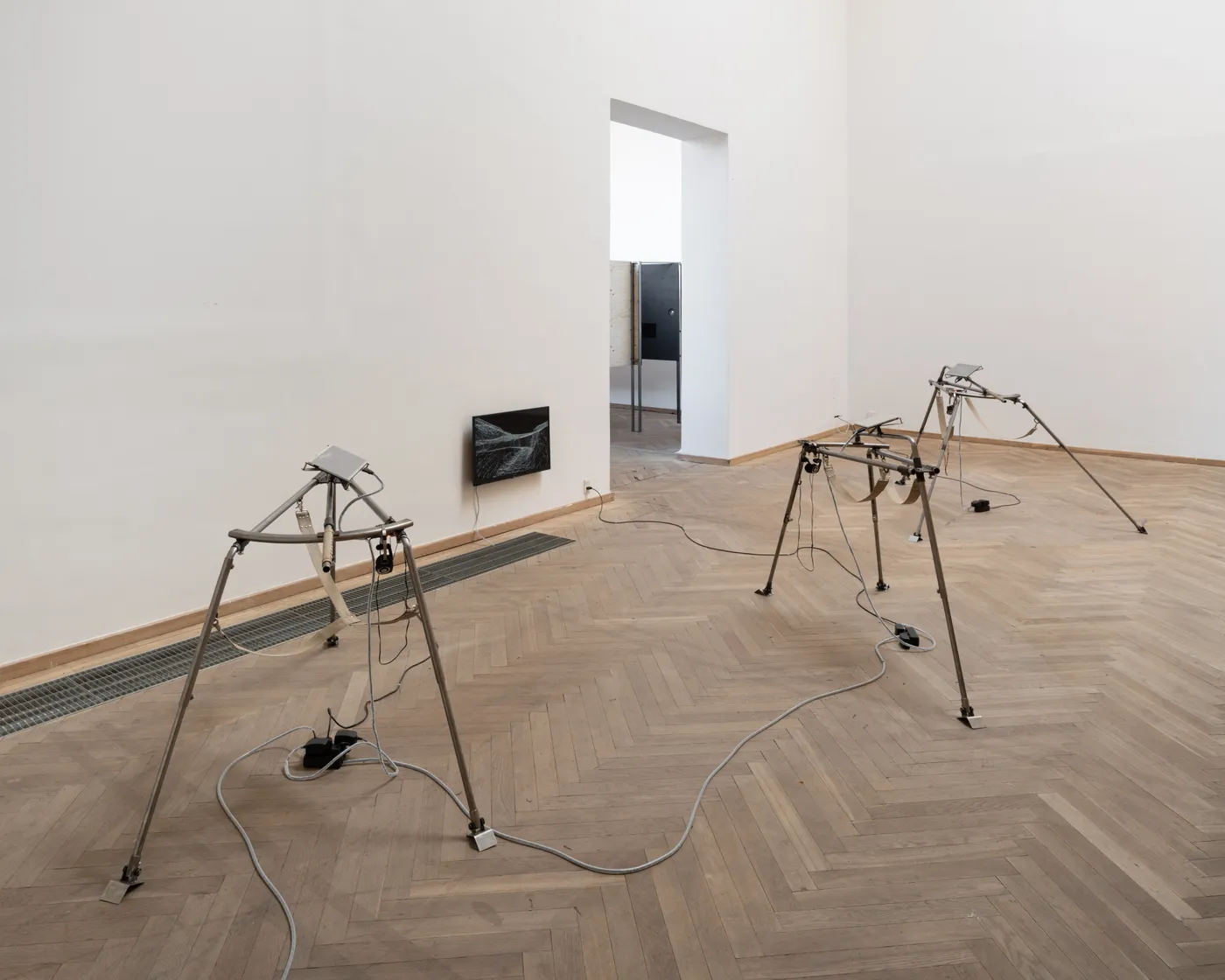 Two steel tripods with small screens in a gallery with a herringbone floor, a third screen on the wall