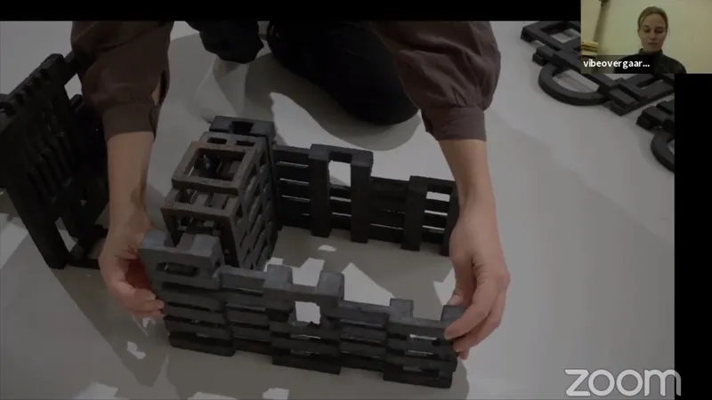 Video call: hands building a structure from dark cast blocks, Vibe Overgaard in a small window
